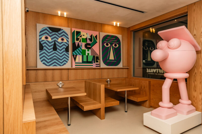 Wooden booths at Softville NYC with tables arranged in front of three framed abstract art prints and a large pink cartoon-style sculpture.