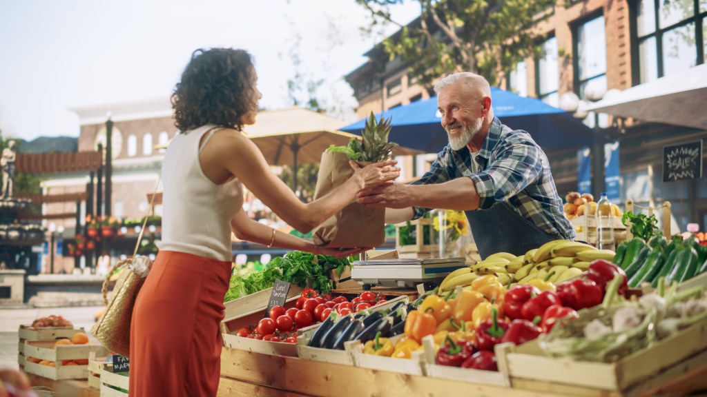 A smiling vendor handing a bag of fresh produce to a customer at a lively outdoor farmers market.