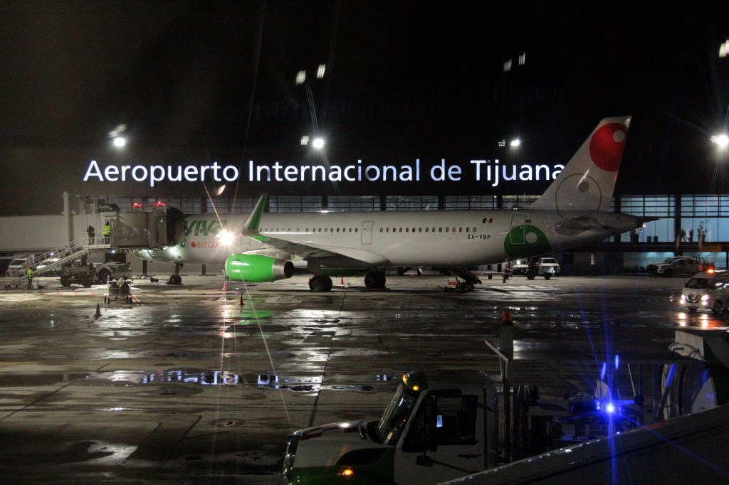 An airliner parked on the tarmac at Tijuana International Airport at night, with the illuminated terminal sign visible in the background.