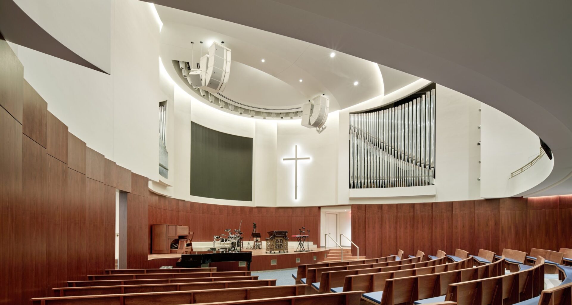 Modern church interior with curved wooden pews, a white illuminated cross on the wall, an organ, and musical instruments on stage under bright lighting—echoing the contemporary design sensibilities of the Lucas Museum of Narrative Art.