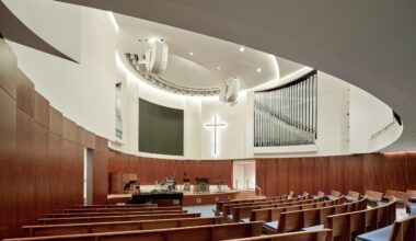 Modern church interior with curved wooden pews, a white illuminated cross on the wall, an organ, and musical instruments on stage under bright lighting—echoing the contemporary design sensibilities of the Lucas Museum of Narrative Art.