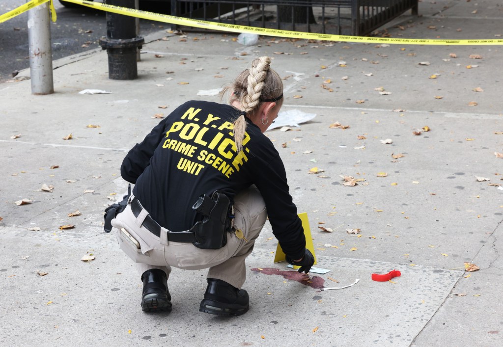 An NYPD Crime Scene Unit investigator crouches to examine a spot on the sidewalk surrounded by yellow police tape.