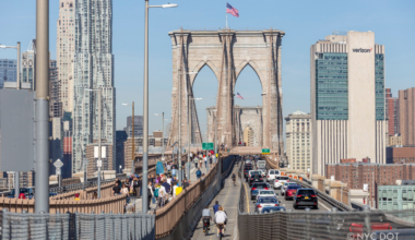 Brooklyn Bridge in New York City on a sunny day, with cyclists and pedestrians crossing the bridge and traffic traveling alongside them. The bridge towers and Manhattan skyline are visible in the background.