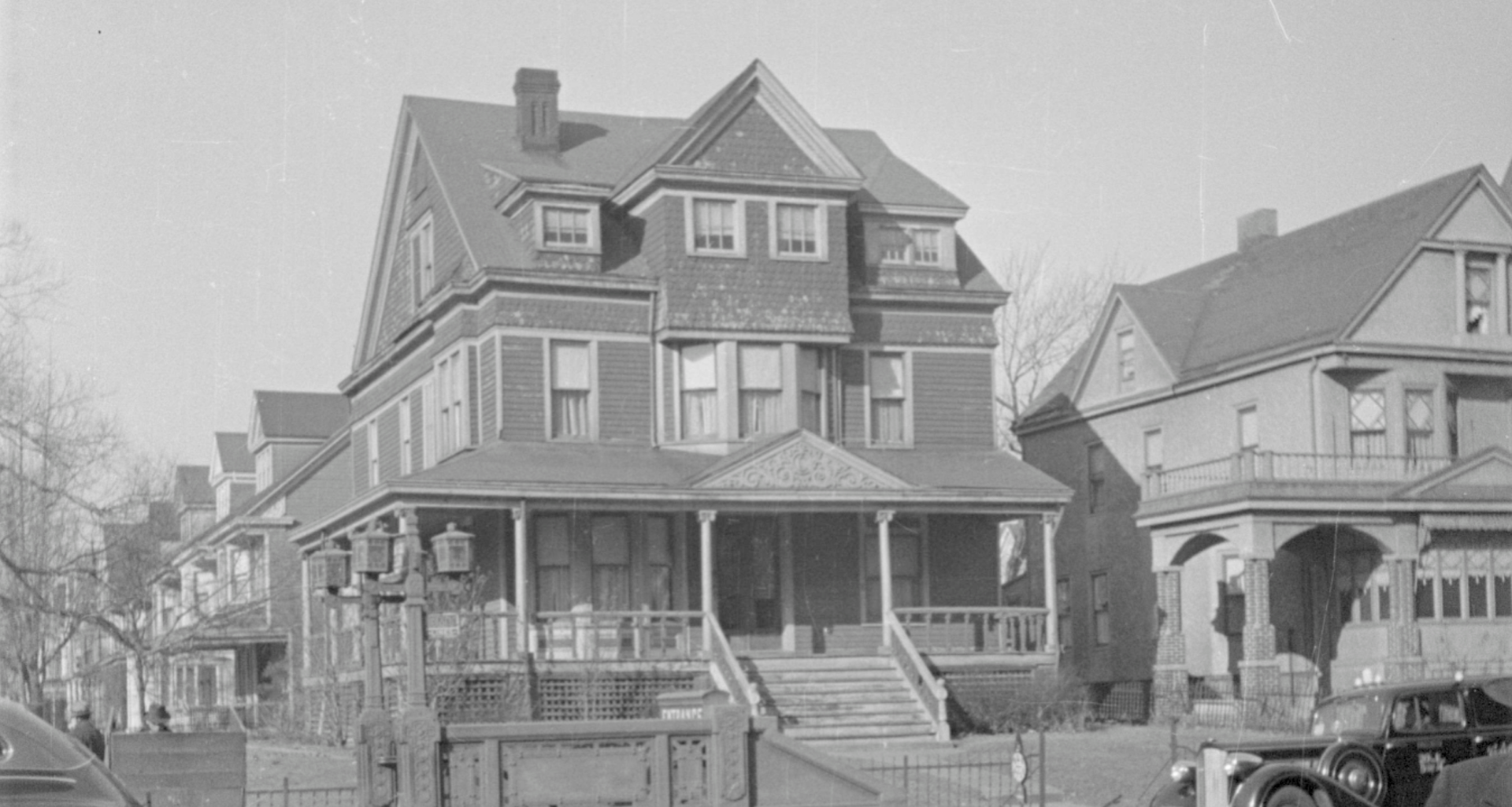 A large, historic Victorian-style house with ornate details and a wraparound porch, situated next to a smaller house. Several trees are in the background, and a vintage car is parked on the street in front.
