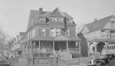 A large, historic Victorian-style house with ornate details and a wraparound porch, situated next to a smaller house. Several trees are in the background, and a vintage car is parked on the street in front.