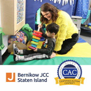 Woman helps a seated child play with colorful blocks in a children’s activity room beside Bernikow JCC and Certified Autism Center logos.
