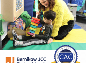 Woman helps a seated child play with colorful blocks in a children’s activity room beside Bernikow JCC and Certified Autism Center logos.
