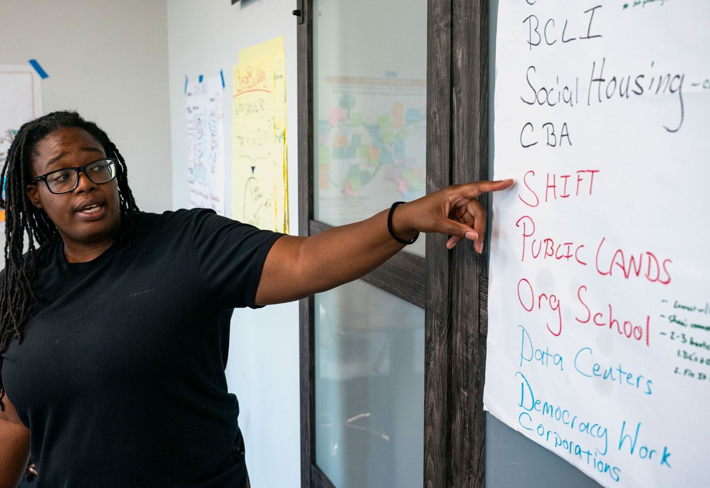 Odessa Kelly, a Black woman in a T-shirt and glasses, points at a hand-lettered flip chart listing organizing priorities, among them social housing, public lands and data centers.