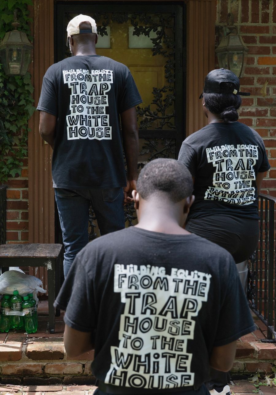 Three Black volunteers, seen from behind, walk up the brick steps to a house's front door. Each wears a black T-shirt printed "Building equity from the trap house to the White House."