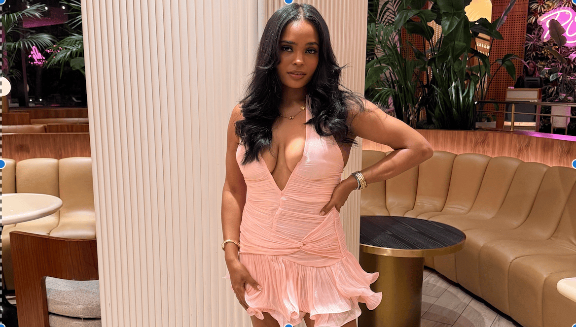 Woman in a peach-colored pleated wrap dress standing in a modern lounge with beige seating, plants, and gold accents in the background.