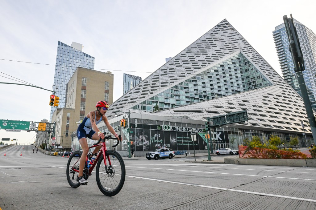 A triathlete in a red helmet rides a bicycle on the West Side Highway during the Big Apple Triathlon, with the pyramidal VIA 57 West building in the background.