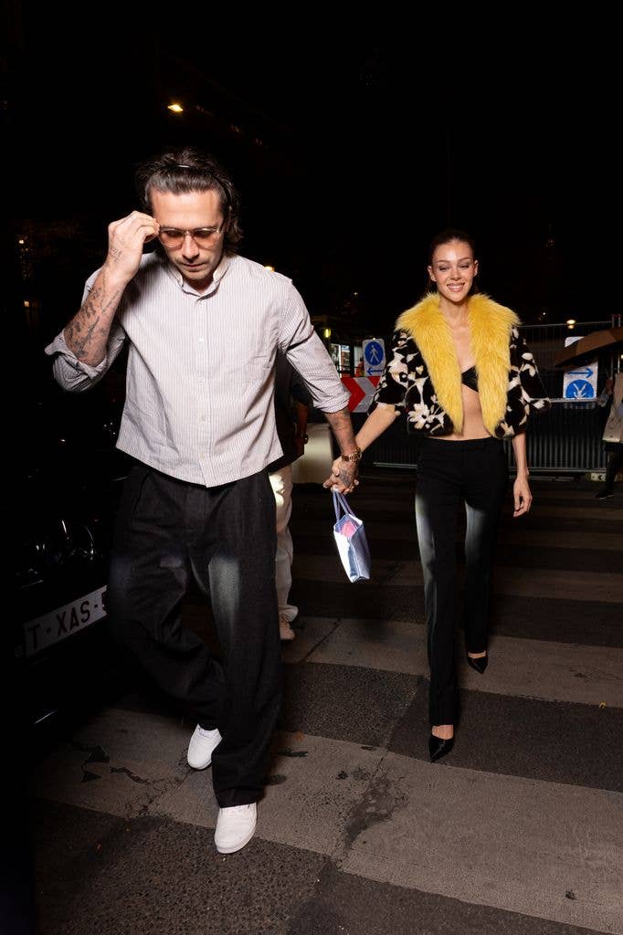 Brooklyn Beckham and Nicola Peltz walking hand in hand at night, he in a striped shirt and she in a yellow fur vest.