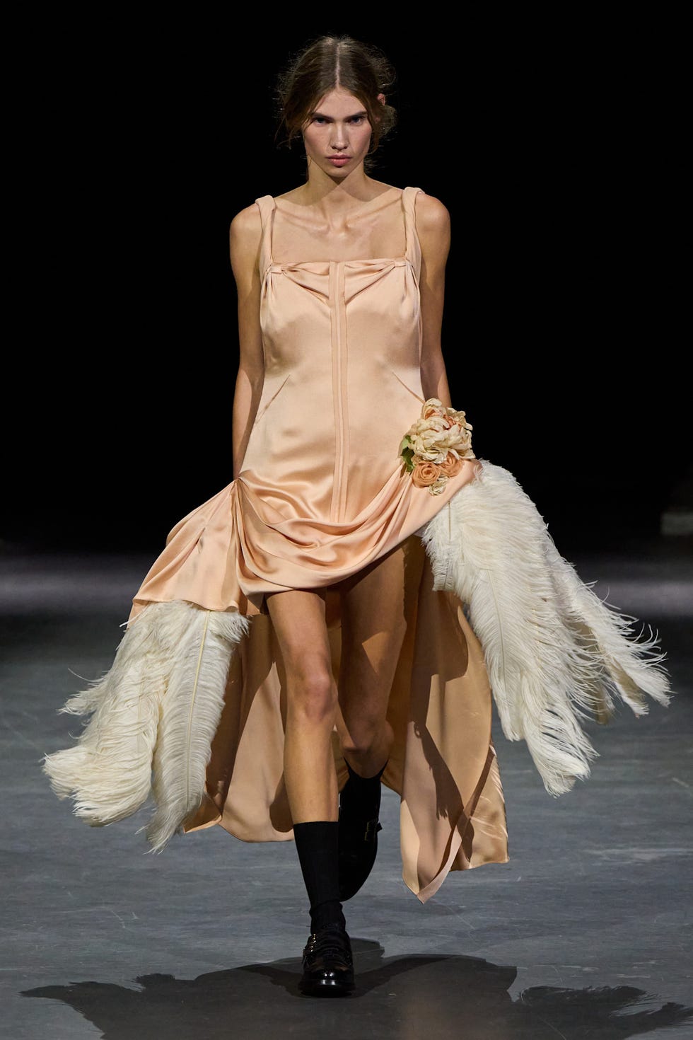 Model wearing a peach satin dress with feather accents and floral embellishments, walking on a runway.