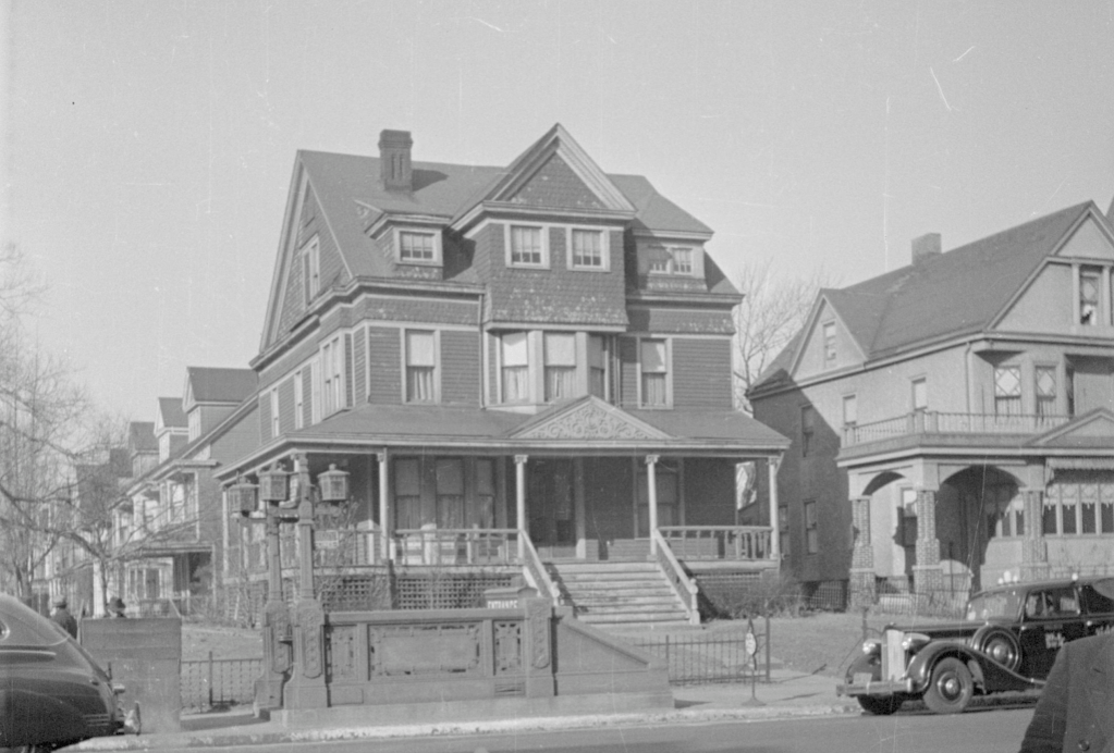 A large, historic Victorian-style house with ornate details and a wraparound porch, situated next to a smaller house. Several trees are in the background, and a vintage car is parked on the street in front.