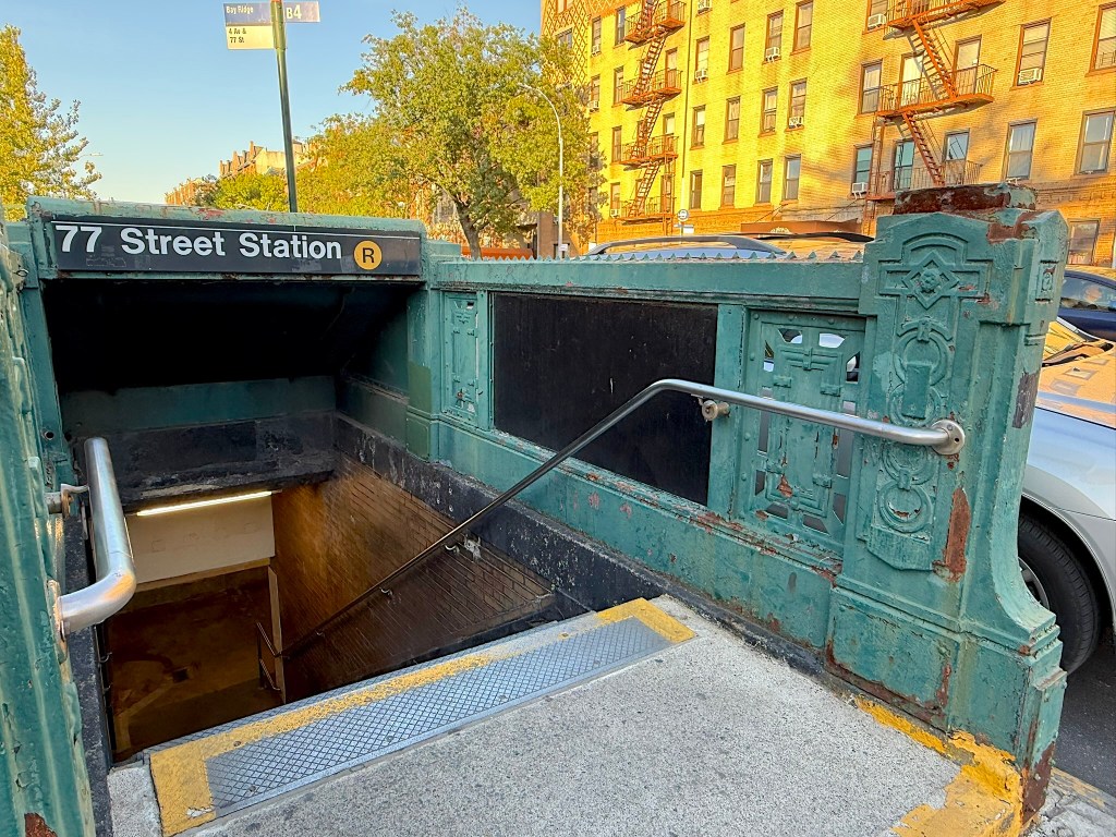 Entrance to 77 Street Station of the New York City subway, featuring worn green ironwork and a staircase leading down, surrounded by urban scenery.