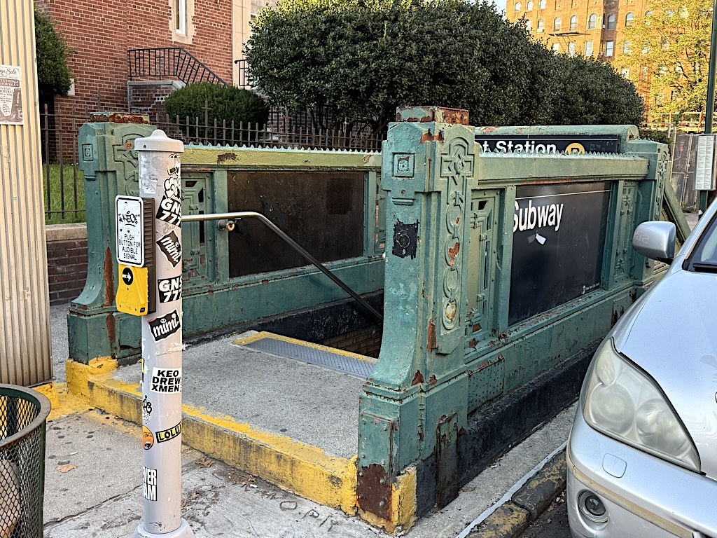 Historic subway entrance with green, weathered metal structure, featuring a 'Subway' sign and adjacent street pole displaying stickers, located near a residential area.