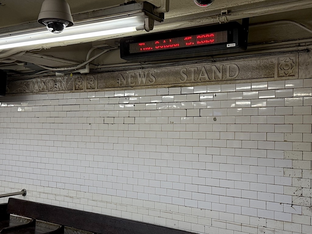 An interior view of a subway station featuring white tiled walls, a sign reading 'NEWS STAND,' and a digital display above showing train information.