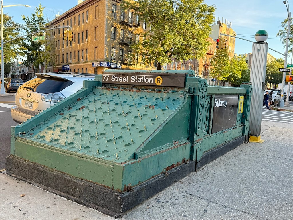 Entrance to 77 Street Station R subway in New York City, featuring a green metal design and nearby urban landscape.