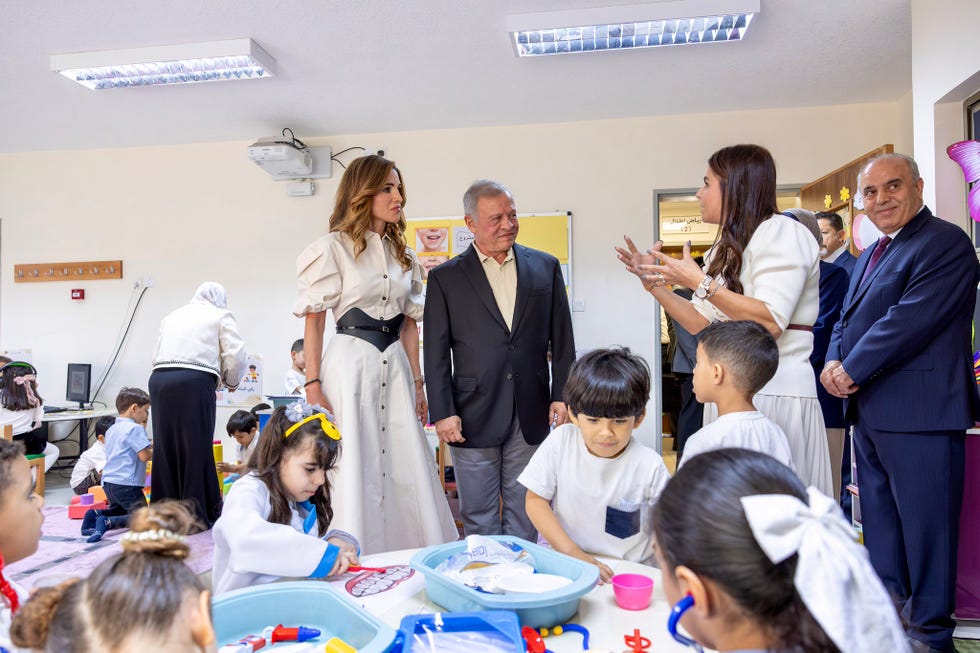 Royal couple visiting a classroom with children engaged in activities while adults converse in the background.