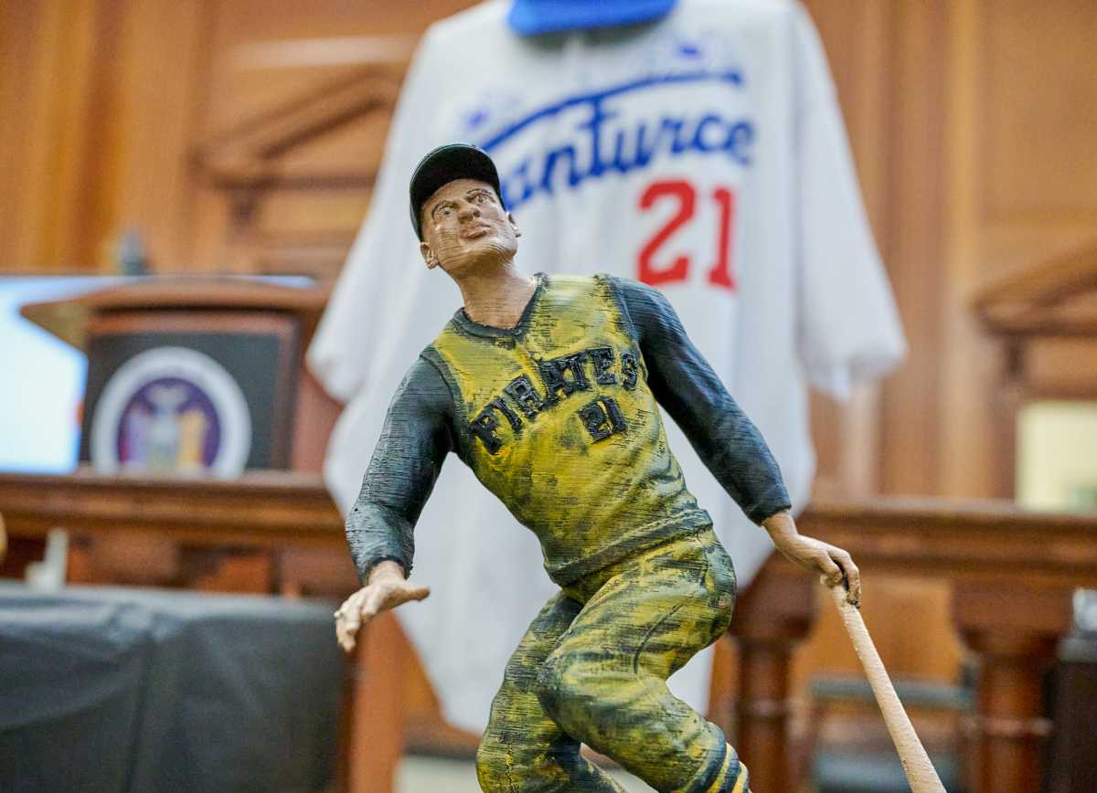 A weathered baseball figurine in a yellow uniform stands in a courtroom, with a white "Pirates 21" jersey displayed behind it.