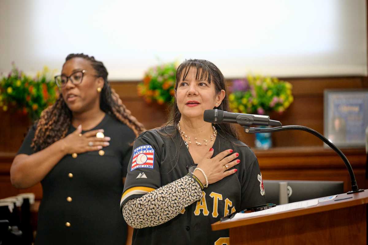 Two individuals stand at a podium, one speaking into a microphone while both place their hands over their hearts.