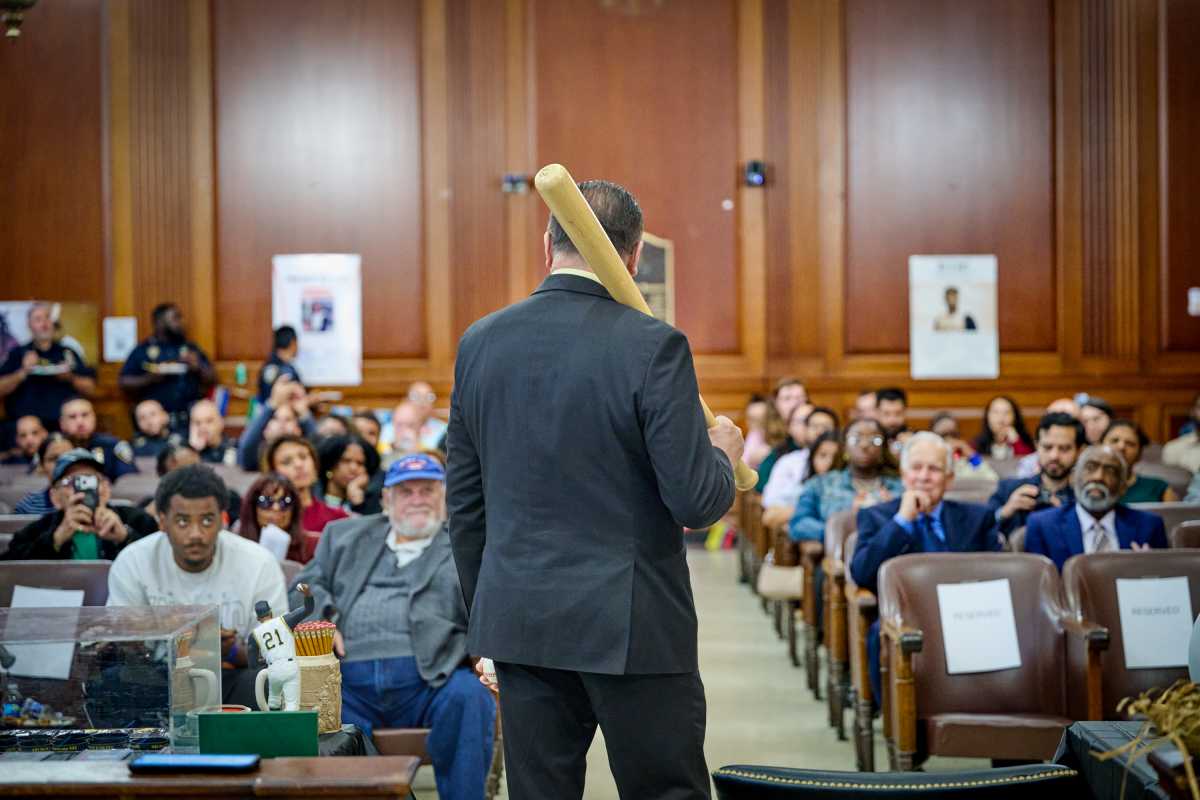 A person in a suit carries a baseball bat over their shoulder while standing in a courtroom filled with spectators.