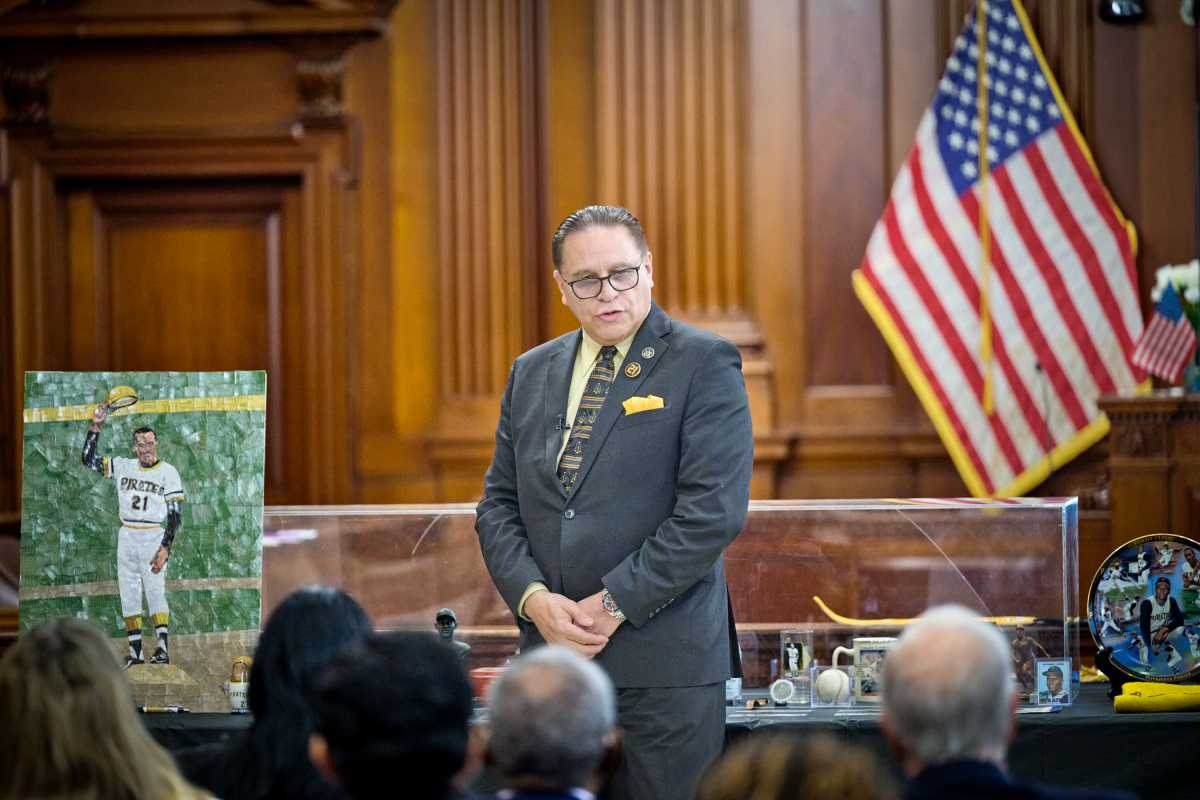 A person in a suit stands before an audience in a formal room, with an American flag and a display case containing memorabilia, including a mosaic portrait of a baseball player.