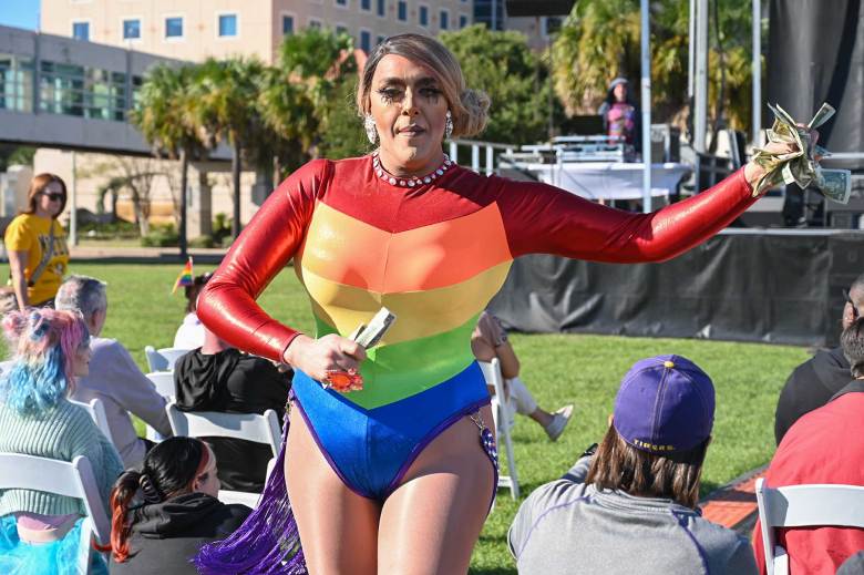 A drag queen wearing a rainbow pride outfit performs among the seated crowd at a daytime festival held outdoors