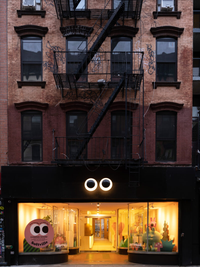 A storefront with a glowing “OO” logo above the entrance and colorful character displays in the windows, set in a brick building with a black fire escape.