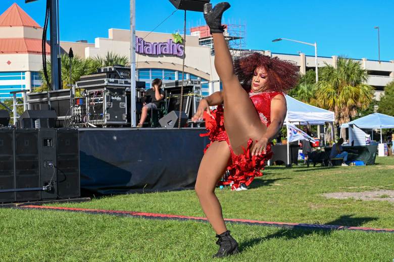 a drag queen in a red sparkly dress kicks her foot into the air while performing at an outside festival