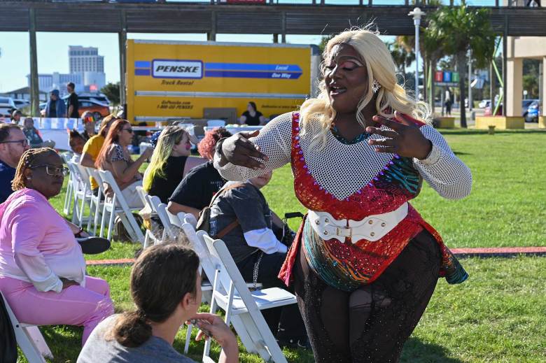 a drag queen wearing a long blonde wig, sparkly red jumpsuit and white belt performs among the seated crowd at an outside festival