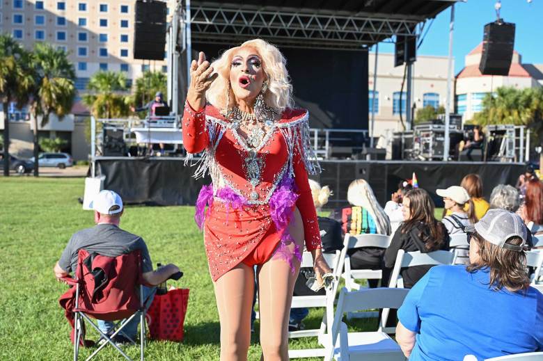 a drag queen with a blonde wig and sparkly red and pink outfit performs for the crowd at an outside festival