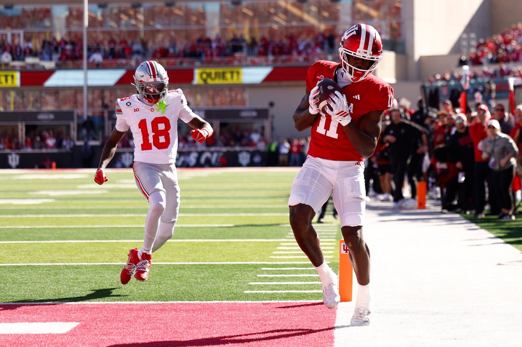 An Indiana football player in a red jersey catches a pass in the end zone while an Ohio State defender in a white jersey pursues him.