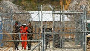 A man with medium-toned skin in an orange prison uniform is transported by guards behind barbed wire fencing.