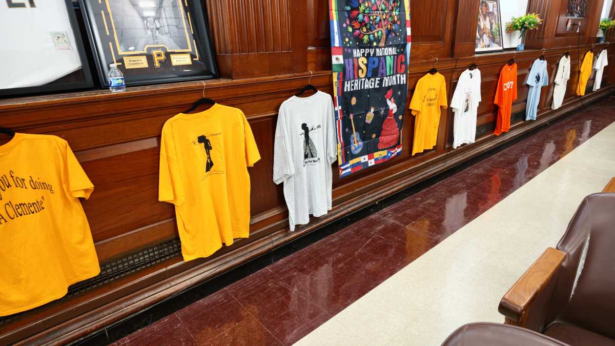 A row of colorful t-shirts hangs on a wooden wall beneath a banner celebrating National Hispanic Heritage Month.
