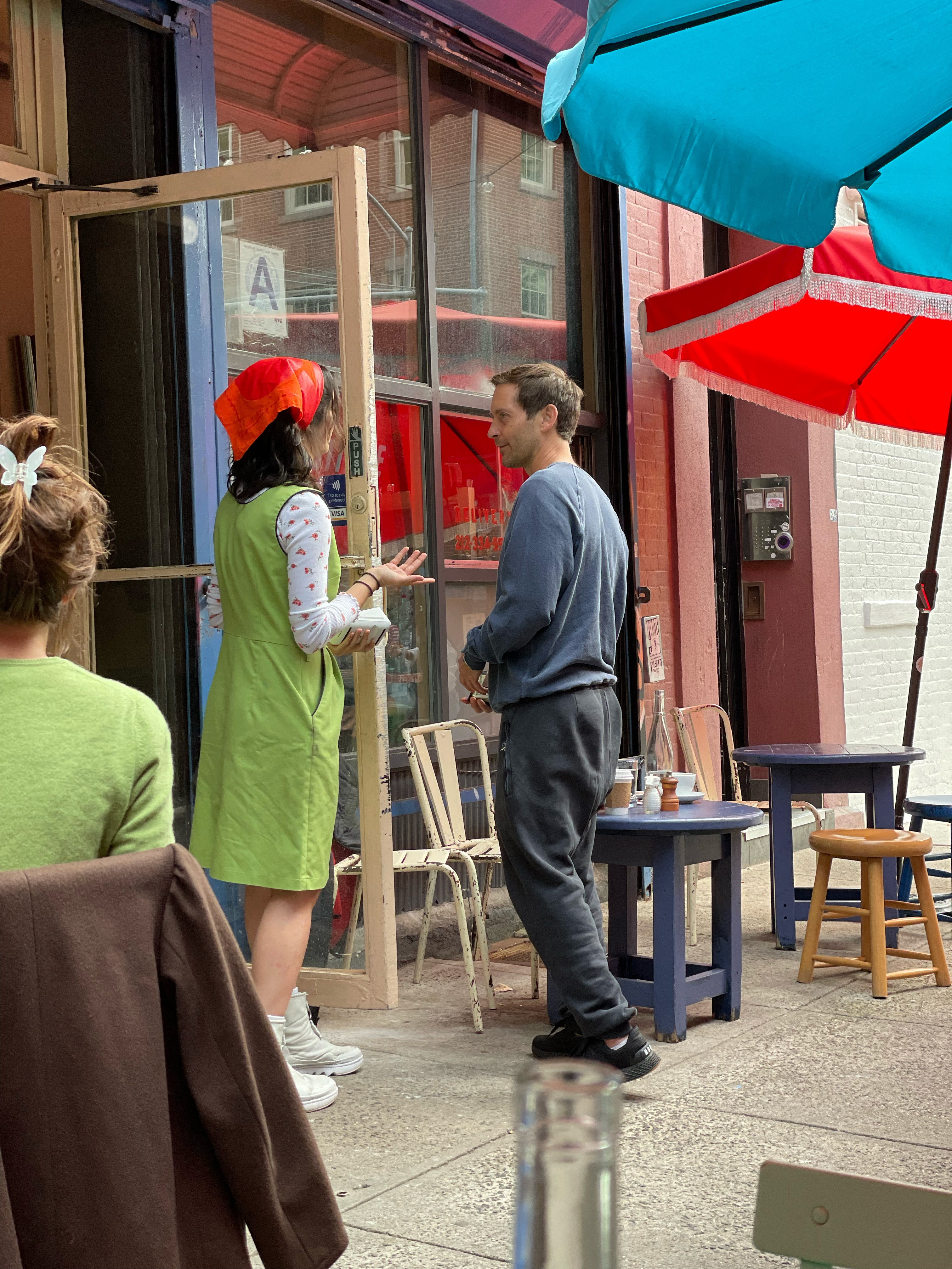 A woman in a green dress and red headscarf talks to a man in a blue shirt outside a cafe with blue and red umbrellas.