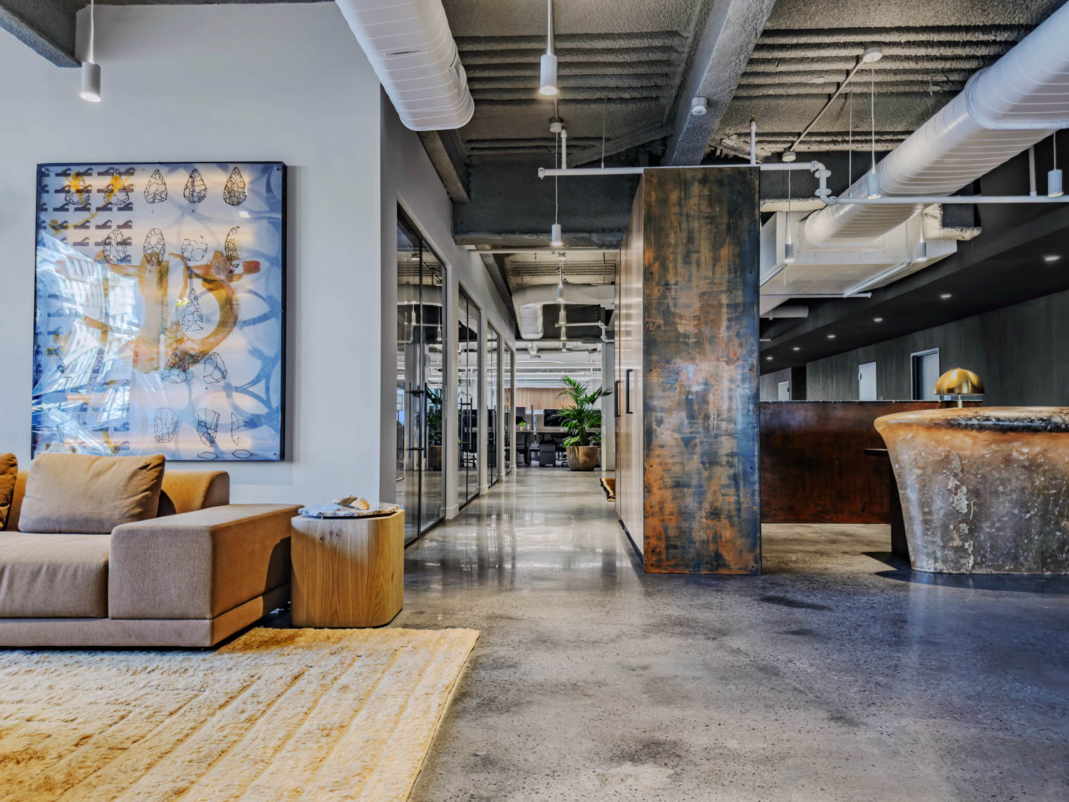 Modern office interior with a tan sofa, concrete floors, and a metal-paneled partition leading to a hallway.