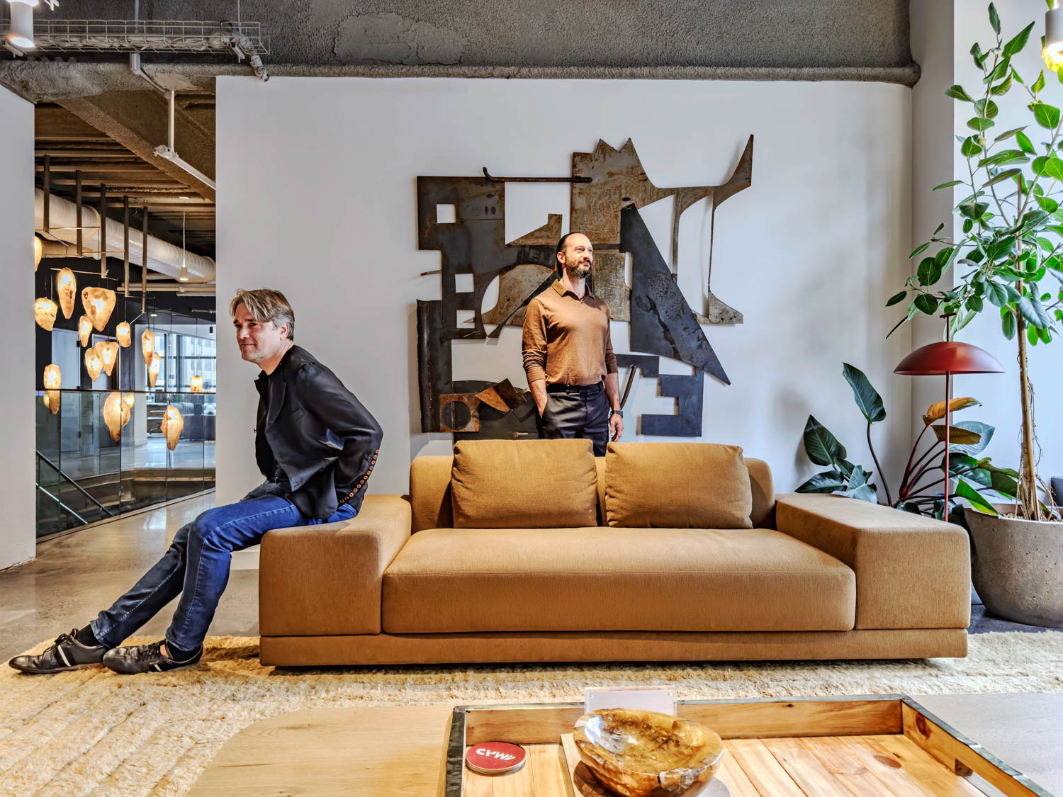 Two men in a modern office lounge featuring a tan sofa and an abstract metal wall sculpture.