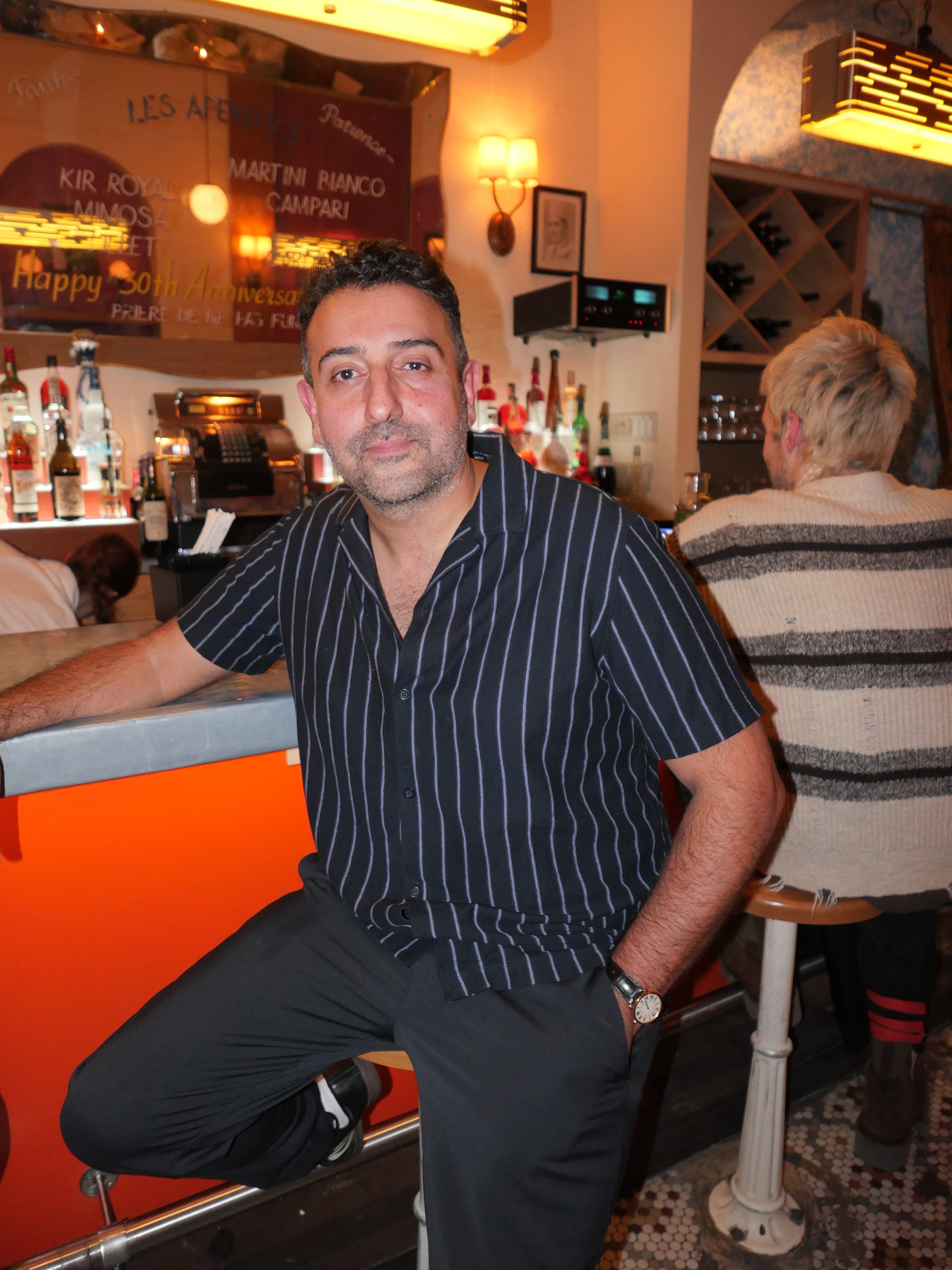 Man with a beard and striped shirt sits at a bright orange bar, another person with blonde hair sits behind him.