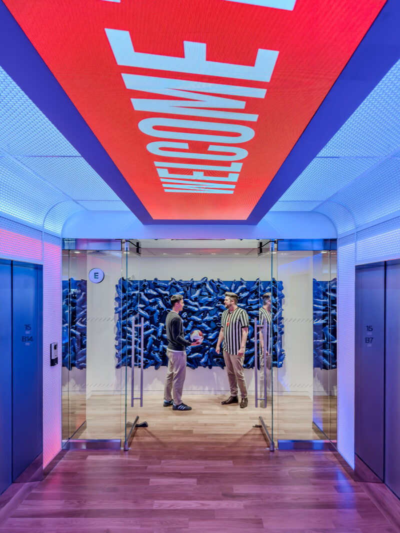 Three people stand and talk in a hallway with glass doors, blue wall art, and a red LED sign on the ceiling reading “WELCOME.” The scene is brightly lit with modern decor.