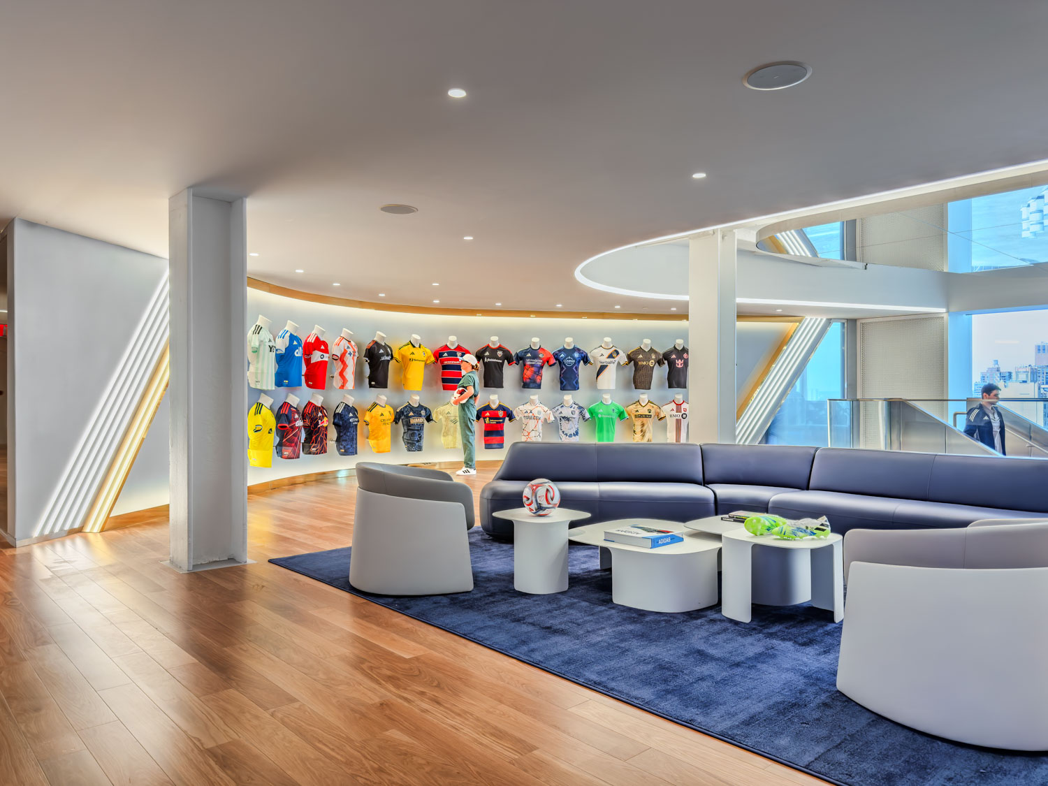 Modern lounge area with curved seating, round tables, and a wall display of various soccer jerseys in a brightly lit room with wooden flooring.