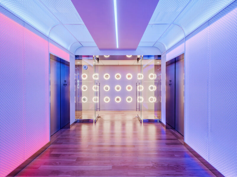 A modern hallway with wooden floors, surrounded by elevators and illuminated by pink, blue, and white LED lighting, leading to a glass wall with circular lights.