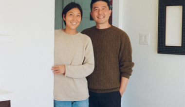 A smiling couple stands in a doorway; the woman wears a beige sweater and jeans, and the man wears a brown sweater and black pants.