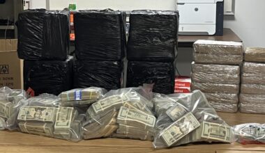 Table in an office holding stacks of black plastic-wrapped packages, several bundles wrapped in tan plastic, and clear bags filled with stacks of cash.