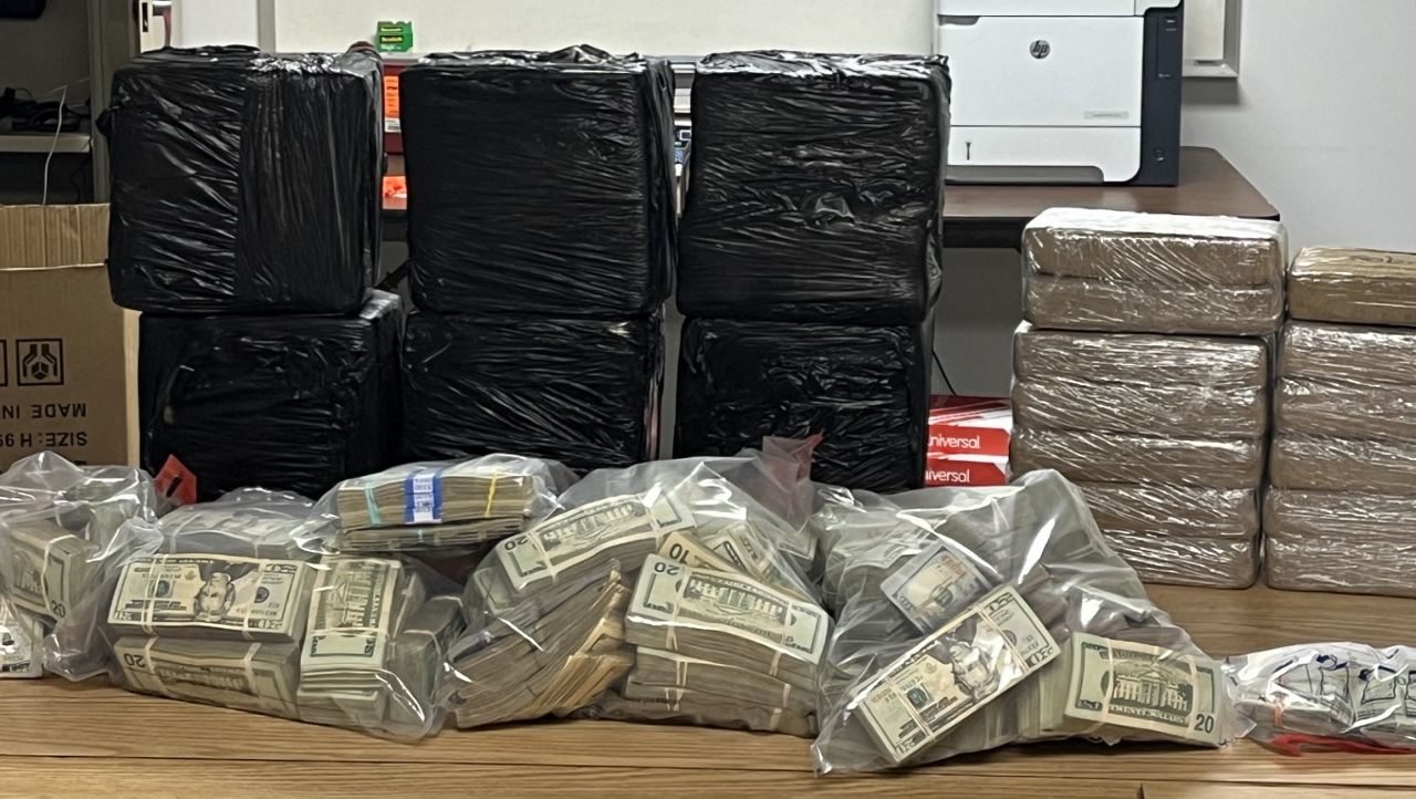 Table in an office holding stacks of black plastic-wrapped packages, several bundles wrapped in tan plastic, and clear bags filled with stacks of cash.