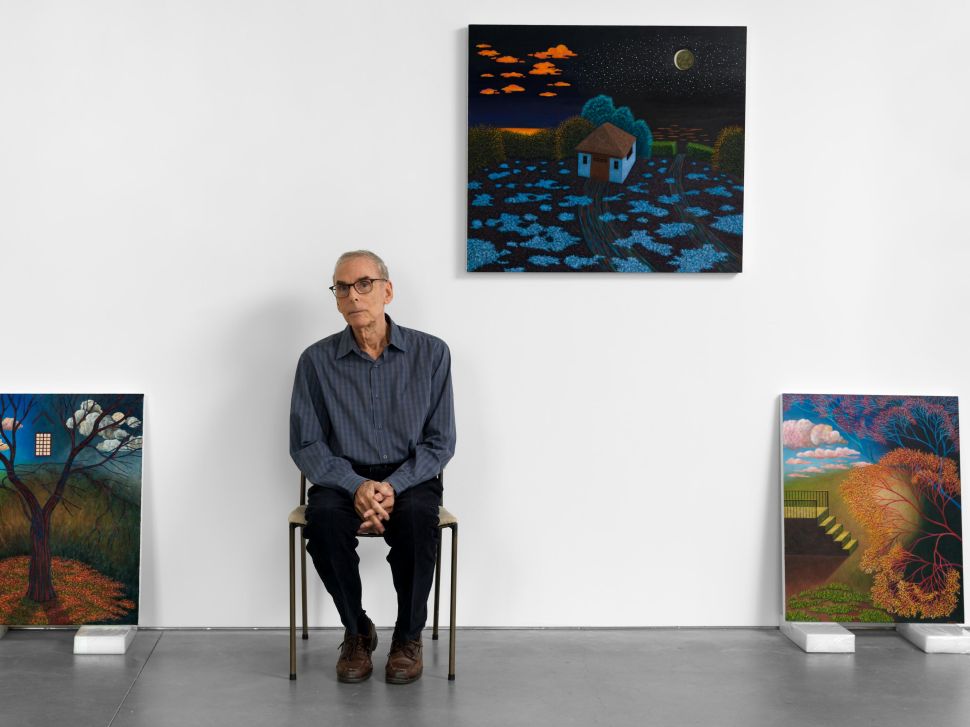 A seated man wearing glasses and a blue shirt poses in a white gallery surrounded by colorful landscape paintings by Scott Kahn.