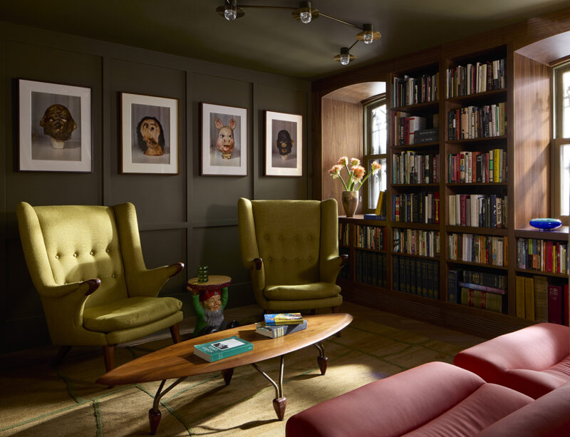 Two lime green wingback chairs face a wooden coffee table in a library with floor-to-ceiling bookshelves and four framed portrait prints on the wall.