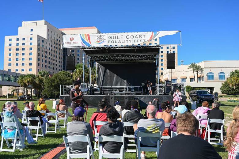 A crowd sits in front of an outdoor stage, a sign reads 'Gulf Coast Equality Fest'