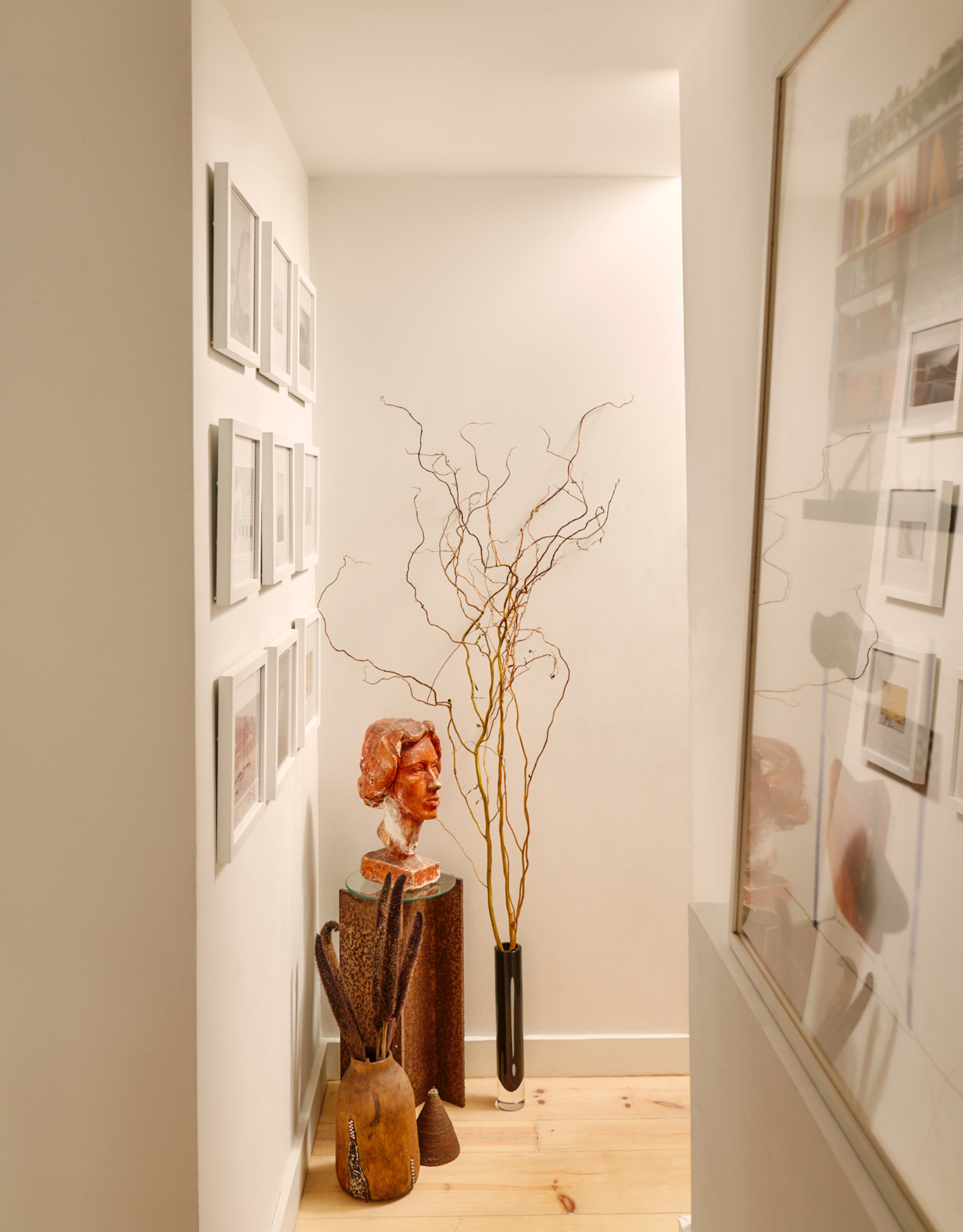 A white corner with a gallery wall, a pedestal with a red bust, a vase of tall twigs, and a ceramic vase