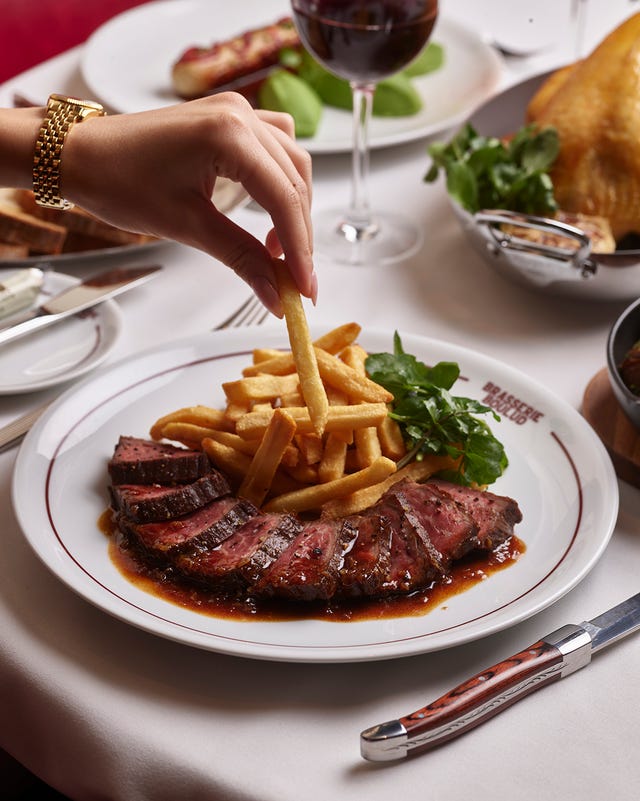 A hand reaching for a French fry next to sliced meat and greens on a dining table with a glass of wine in the background. A hand reaching for a French fry next to sliced meat and greens on a dining table with a glass of wine in the background.